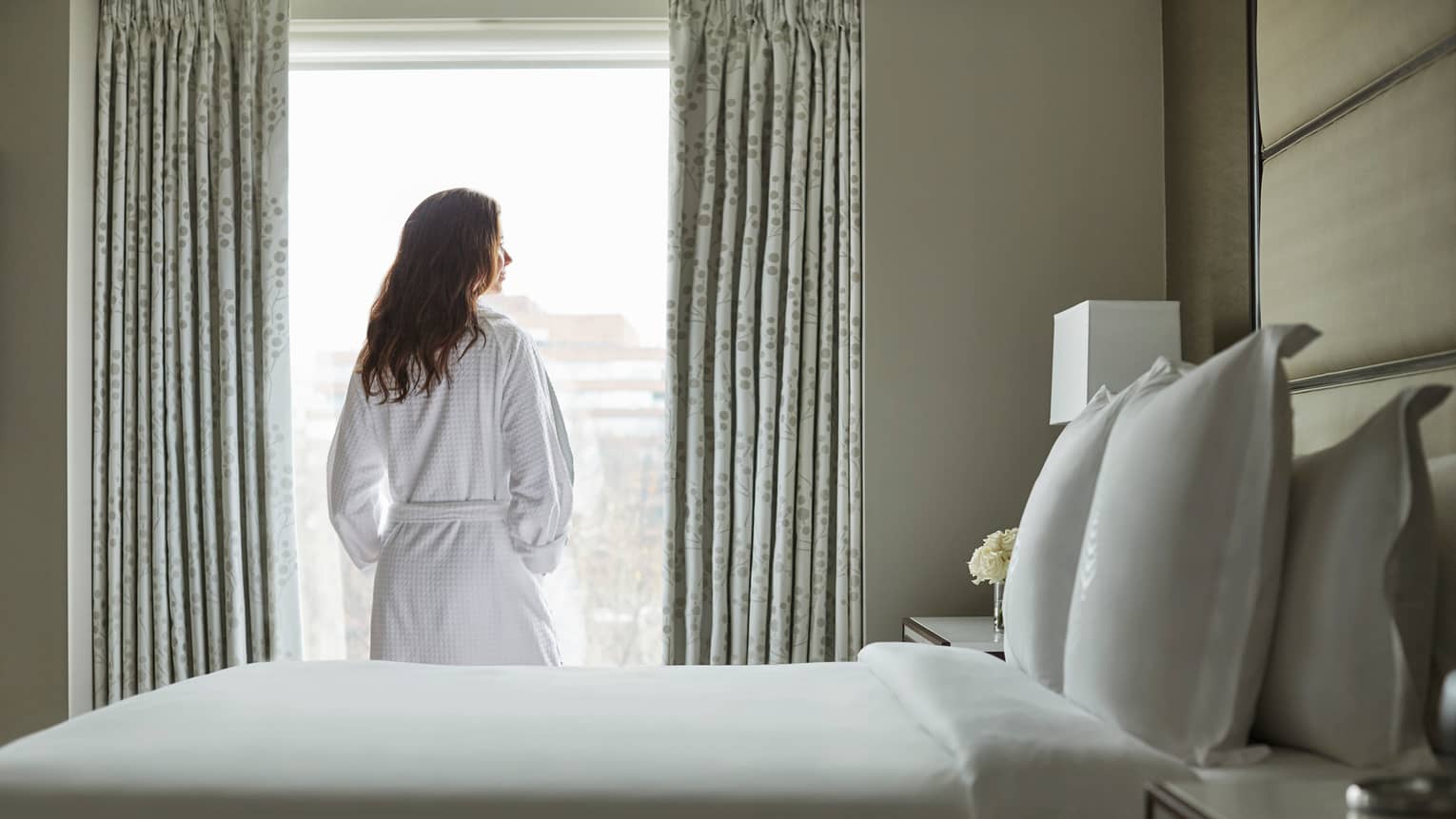 Back of woman in white bathrobe stands at sunny window in front of bed