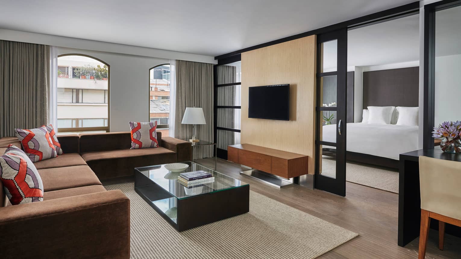 One-Bedroom Suite living room with L-shaped brown sofa, red and grey geometric accent pillows, TV wall divider to bedroom