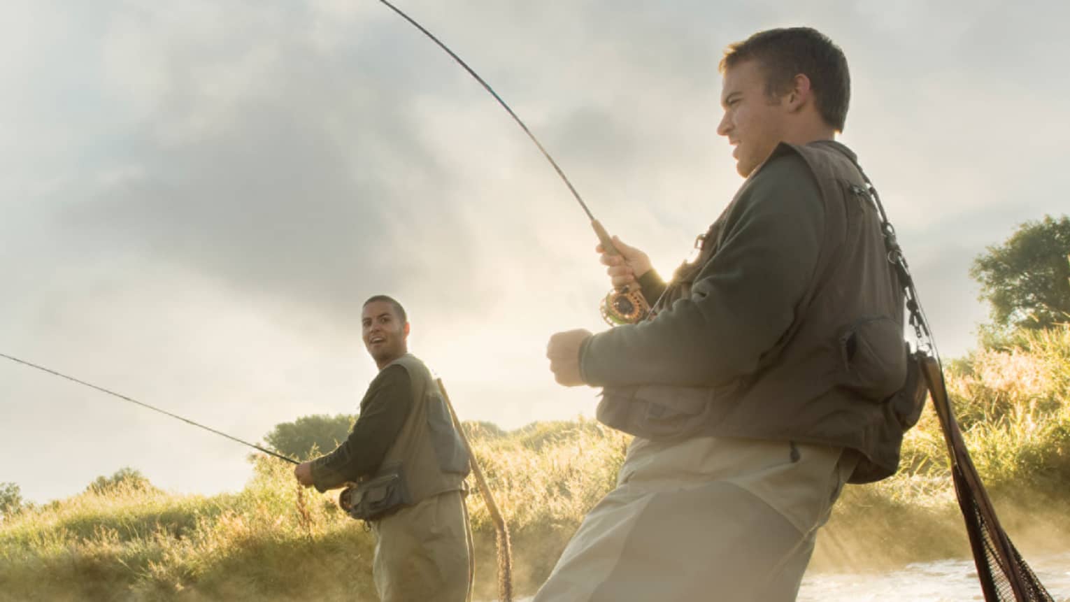 Two people fly fishing in a river, standing knee-deep in the water, surrounded by mist and greenery.