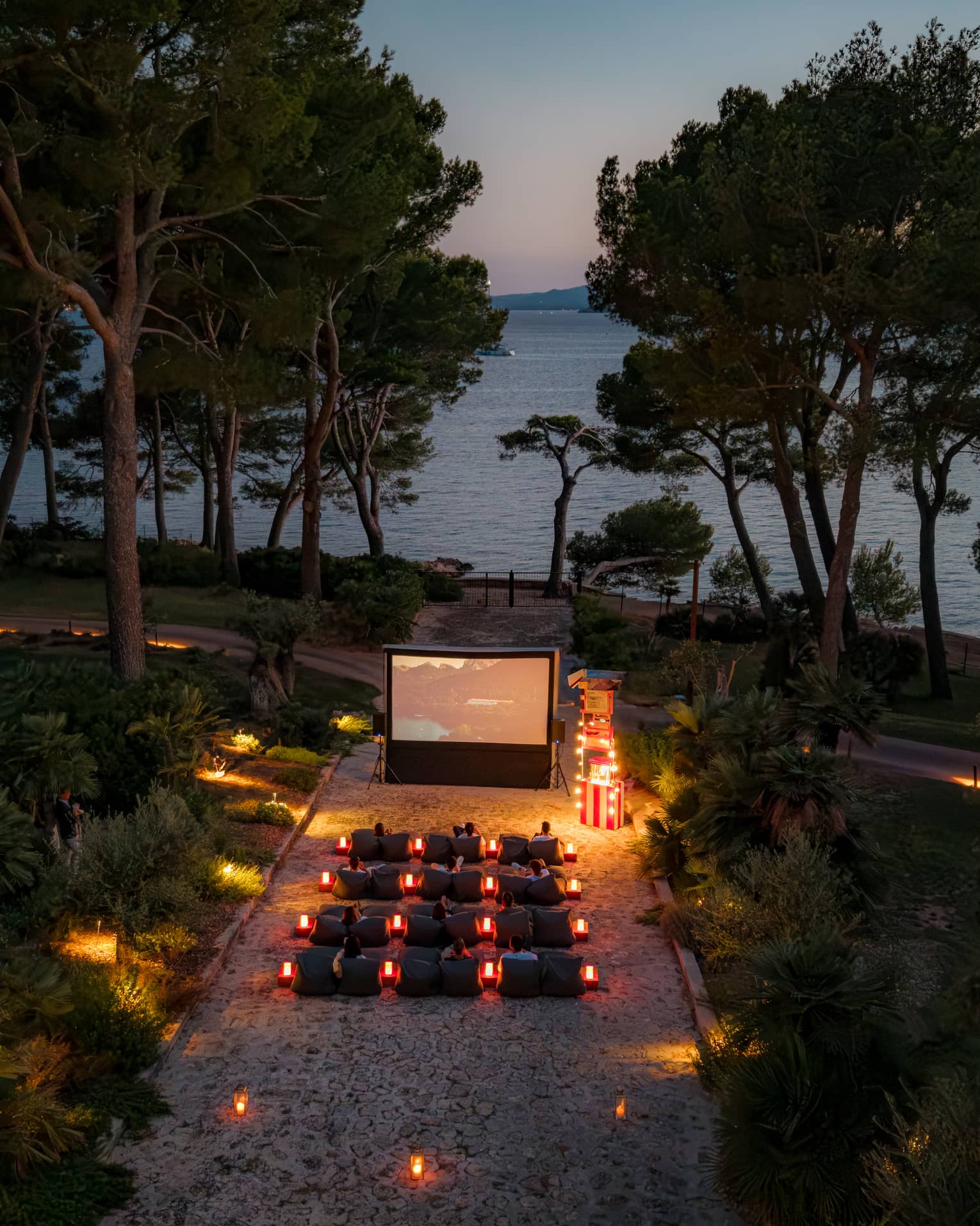 A bird’s-eye view of people seated in rows of chairs in front of a large movie screen in an outdoor setting in the evening, surrounded by trees, with the ocean in the background