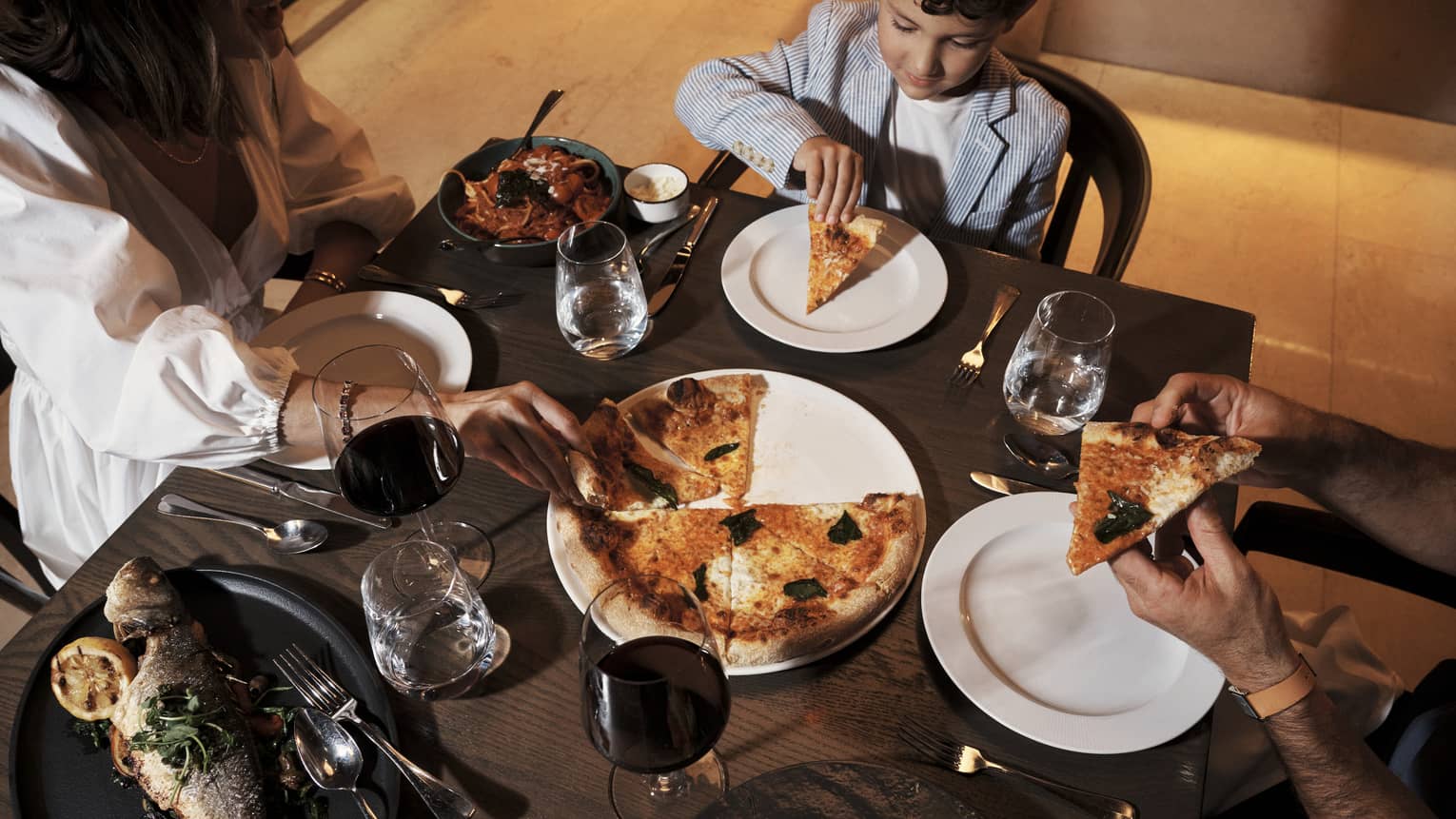 Guests sit at a small dining table set with dinner dishes, including pizza and fish.