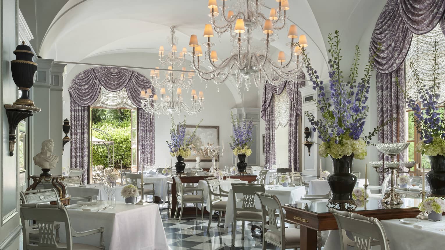 An ornately decorated dining room with warm chandelier lighting and open windows looking out on lush greenery