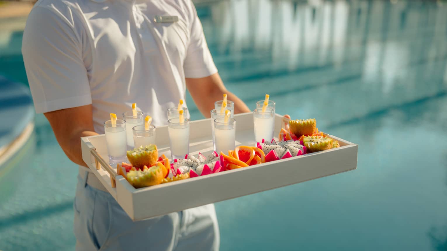 Pool attendant wearing a white short-sleeve polo shirt and light-blue shorts carries a large tray filled with beverages and small plates of colourful fruit and snacks