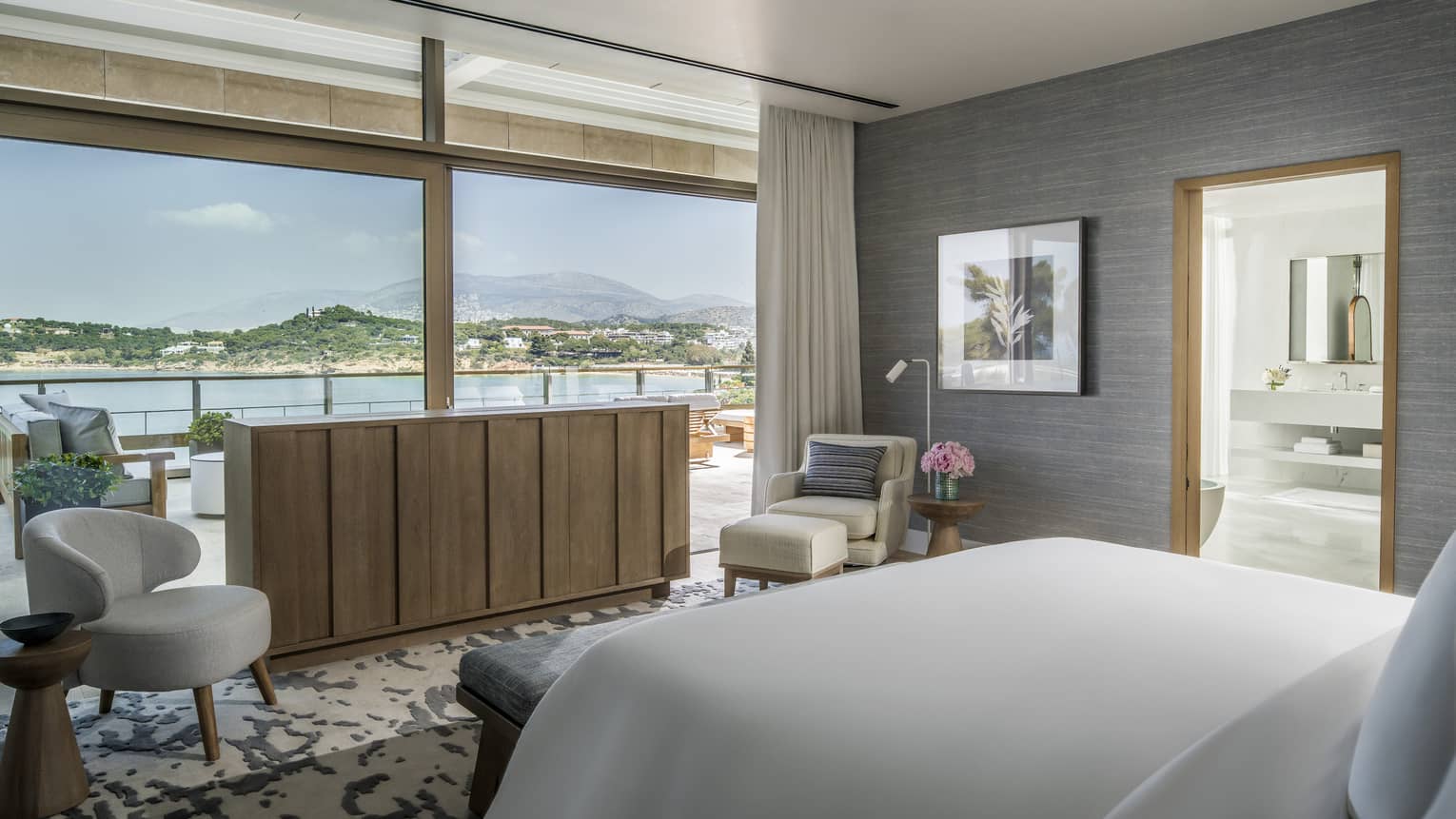 Arion Royal Suite with white King Bed, blonde wood sideboard, grey walls, white chaise and arm chair, doorway to bathroom, floor-to-ceiling windows, ocean views