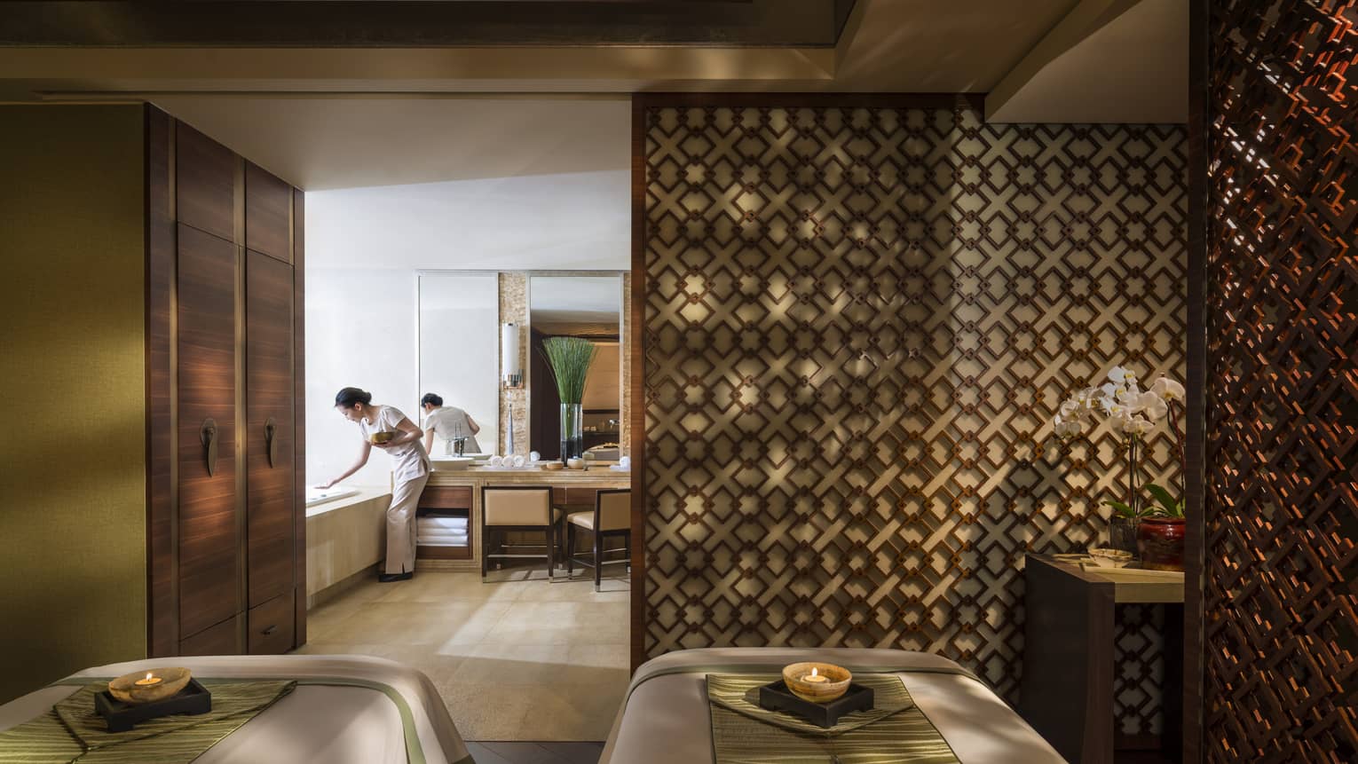 Couples massage beds side-by-side in spa room with candles, wood detail wall, staff preparing bath