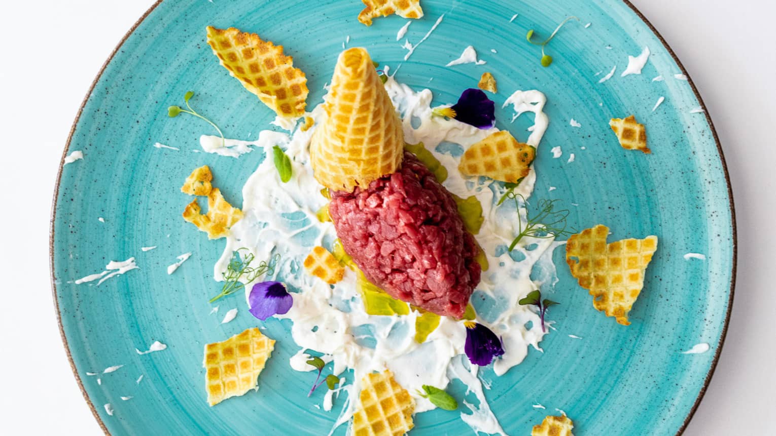 A blue plate with a waffle cone shattered over white cream and a red food.