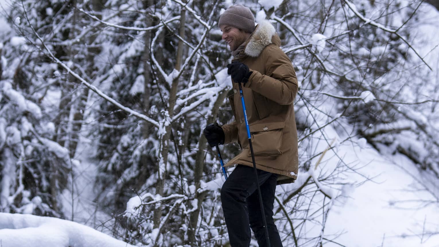 A snowshoer in a knit cap, parka and gloves uses poles to cross fallen logs in a dense, snow-covered forest.