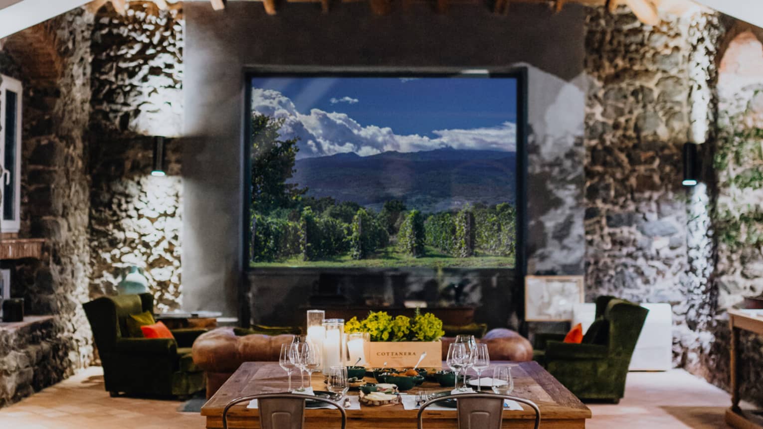 A wooden dining table set with floral arrangements and glassware,  in a room with a window overlooking mountains and a vineyard