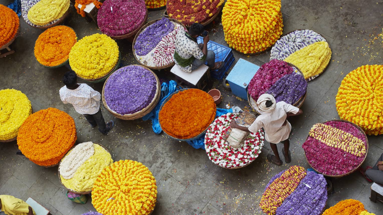 Bird’s-eye view of vendors with chains of vibrant, multicoloured flowers arranged in spirals in large round wicker baskets.