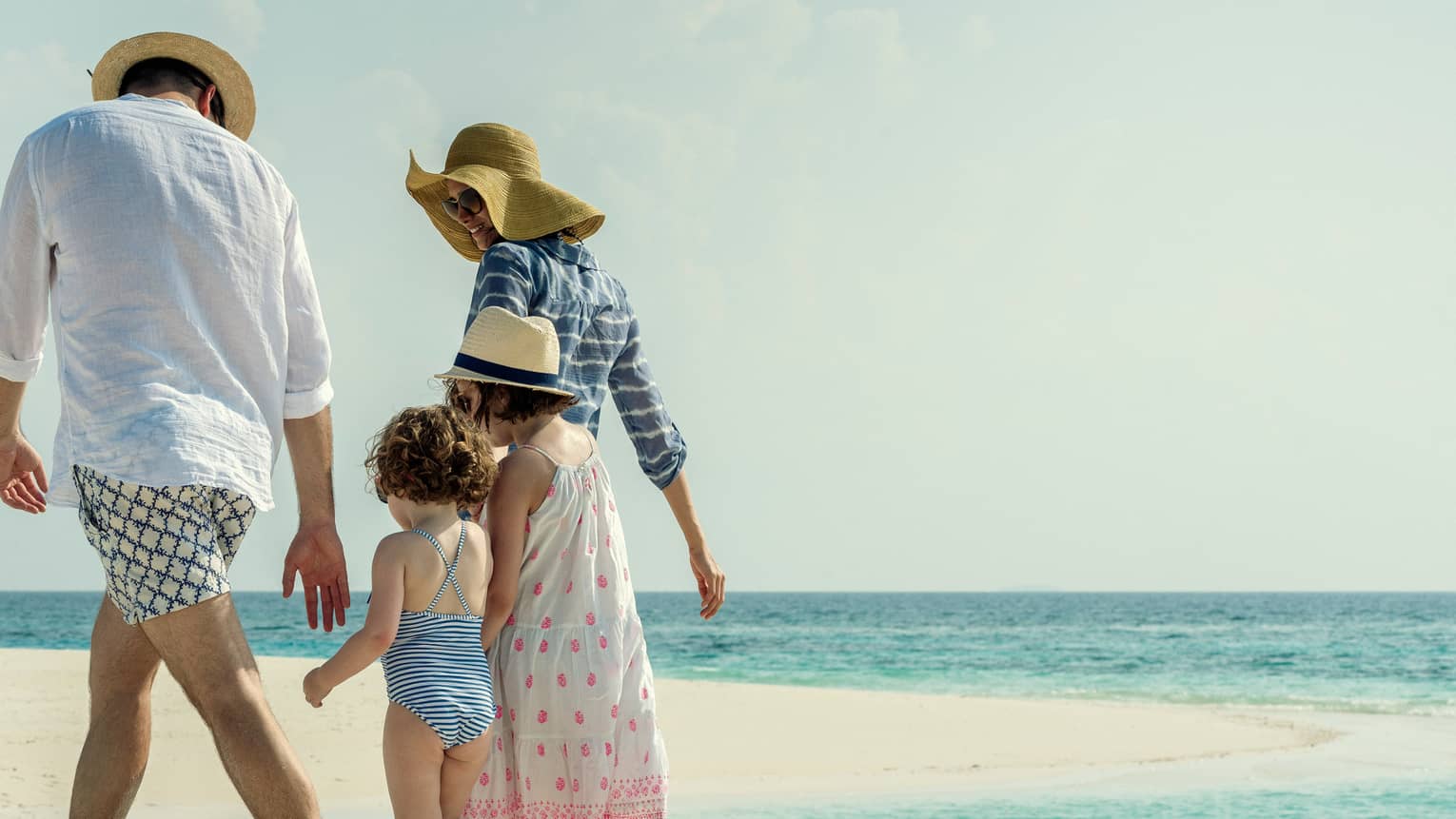Family wearing swimsuits, sundresses and sun hats walk along white sand beach