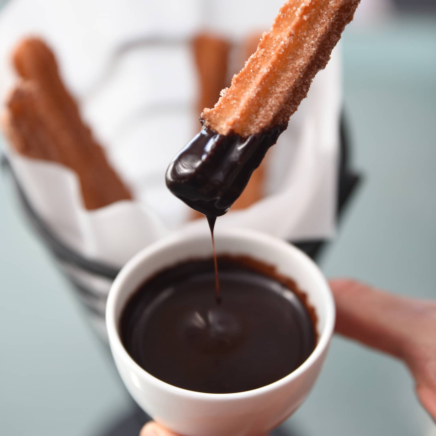 Hand holds small white bowl of melted chocolate, dipping churro into it