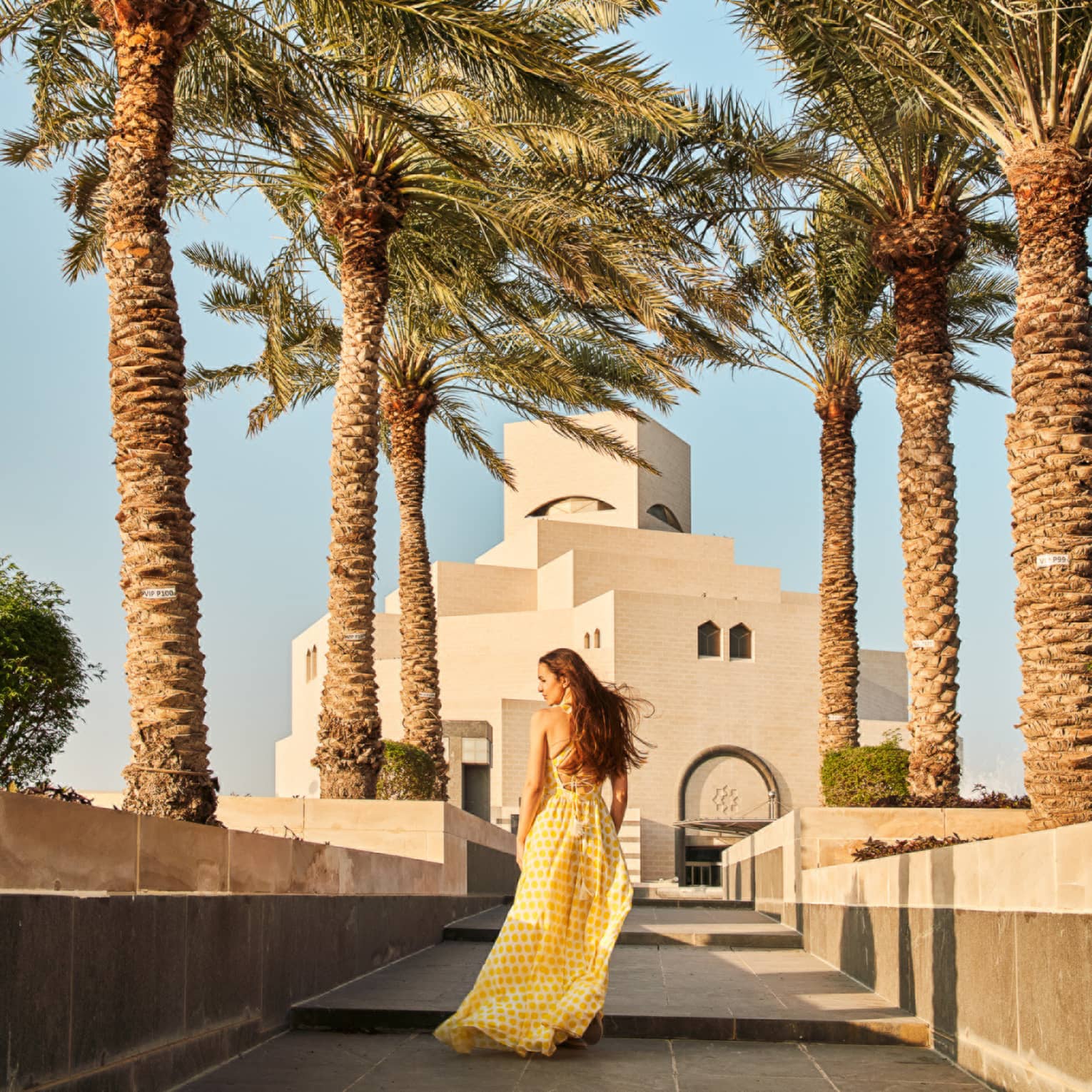 Woman wearing long flowing yellow dress walks along a tiled walkway surrounded by palm trees