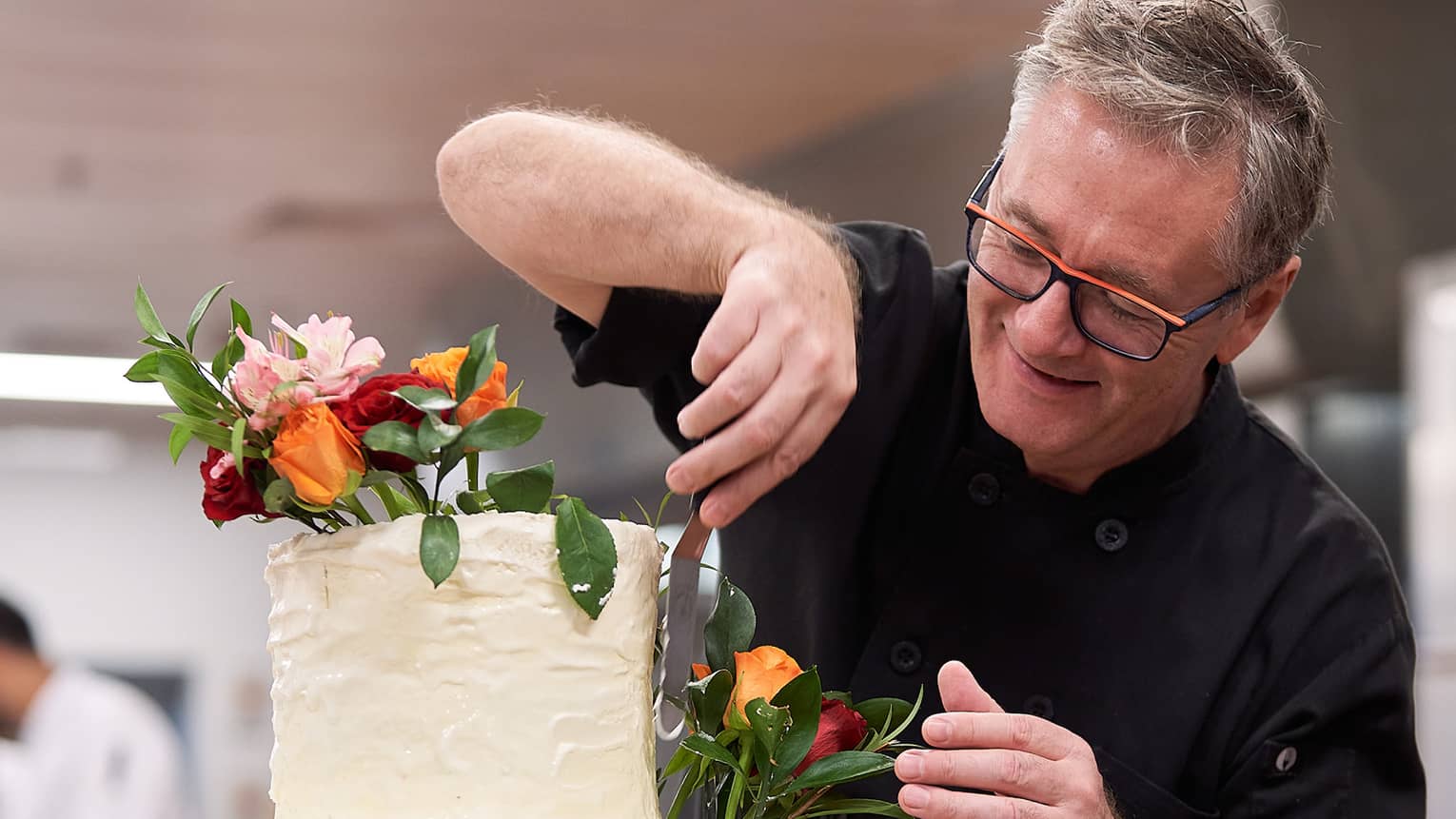 Chef spreads frosting with off-set spatula on top tier of three-tier wedding cake with fresh flowers
