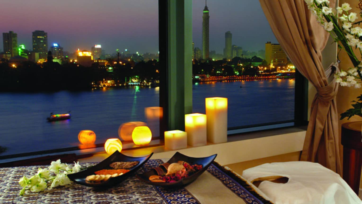 White candles glow along Spa windowsill looking out at river, city lights, small charcuterie platters on table