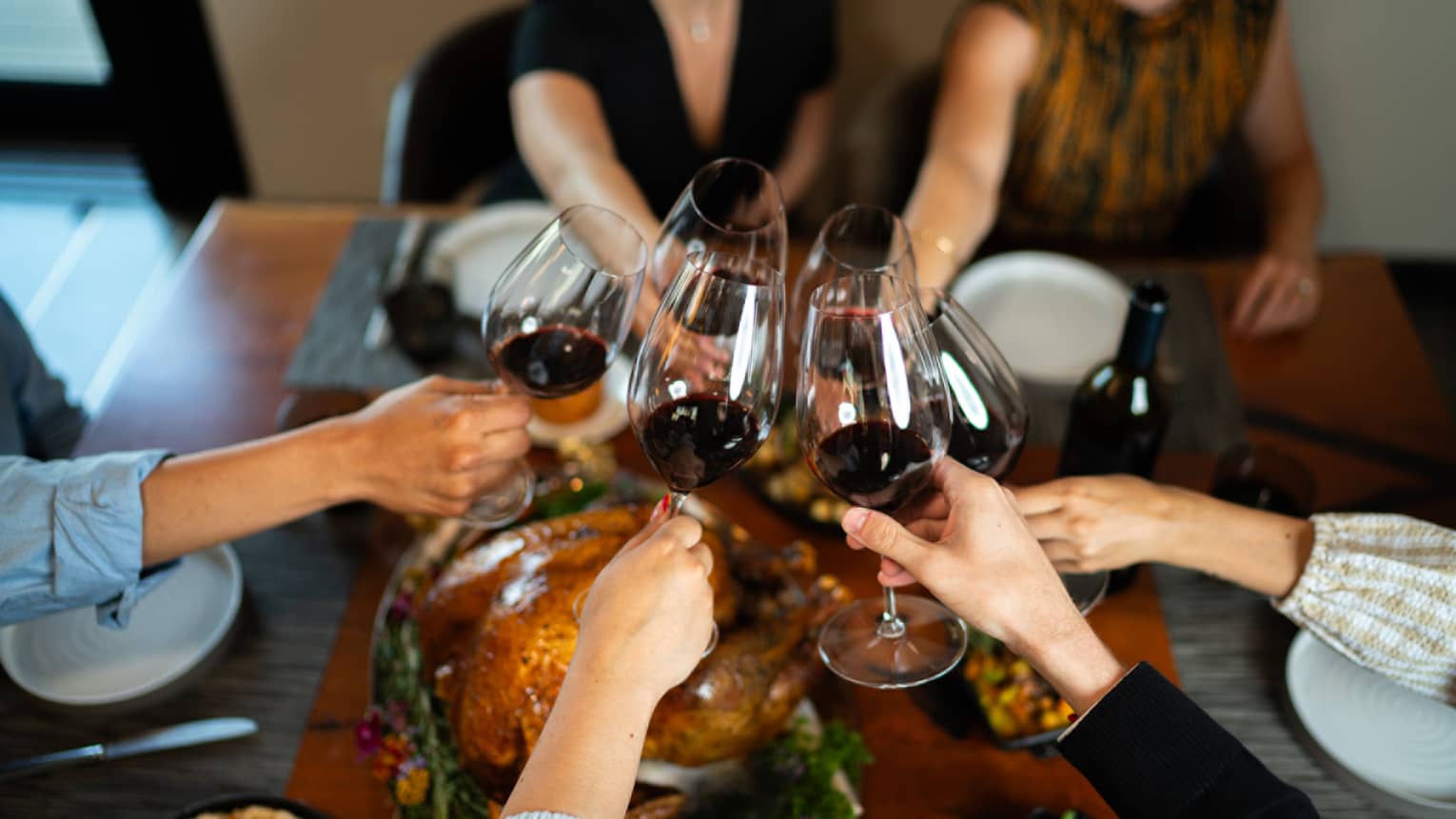 Six arms extend toward the middle of the table to toast over a meal, each holding a glass of red wine