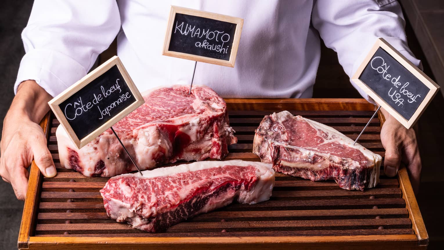 Close-up view of a wood tray with three labelled cuts of well-marbled beef, held by person in white chef's coat.