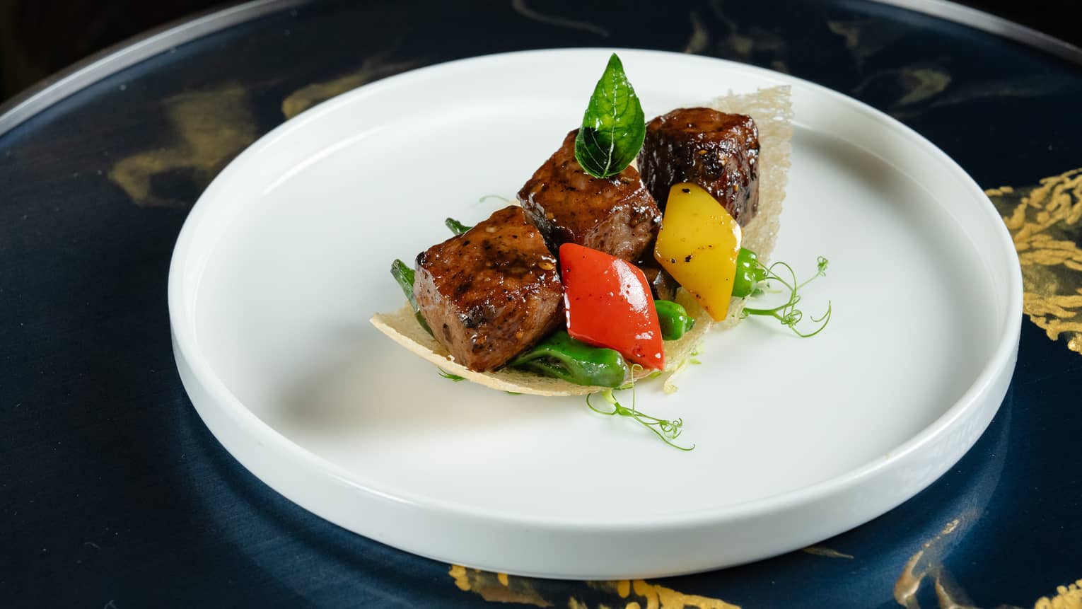 Three cubes of grilled meat with diamond-shaped green and red pepper slices and microgreens, cradled by a curved rice wafer.