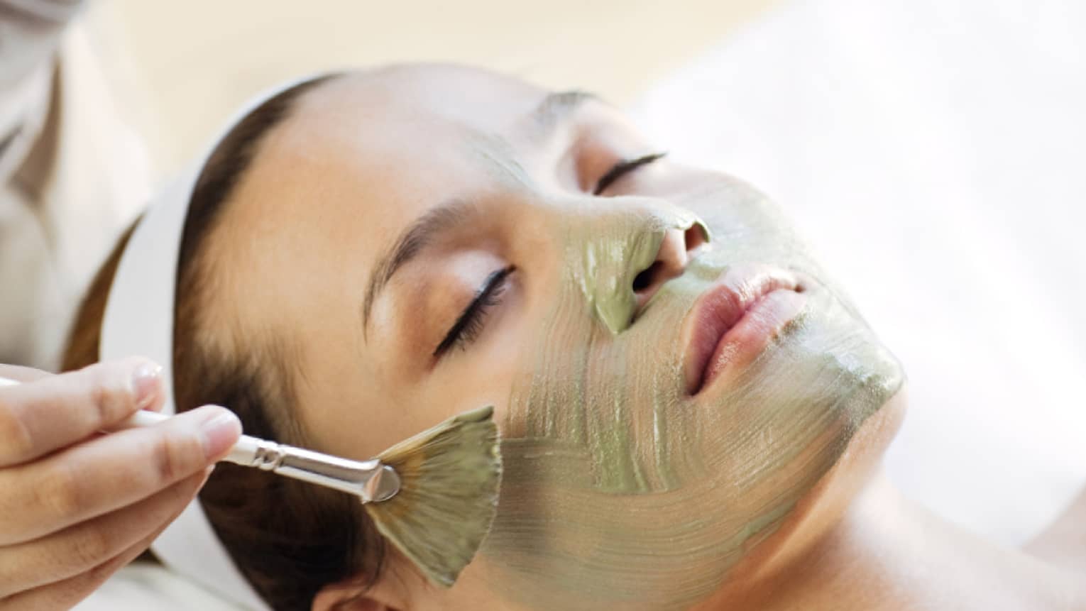 Spa staff holding brush paints green mask on woman's cheeks, chin, nose 