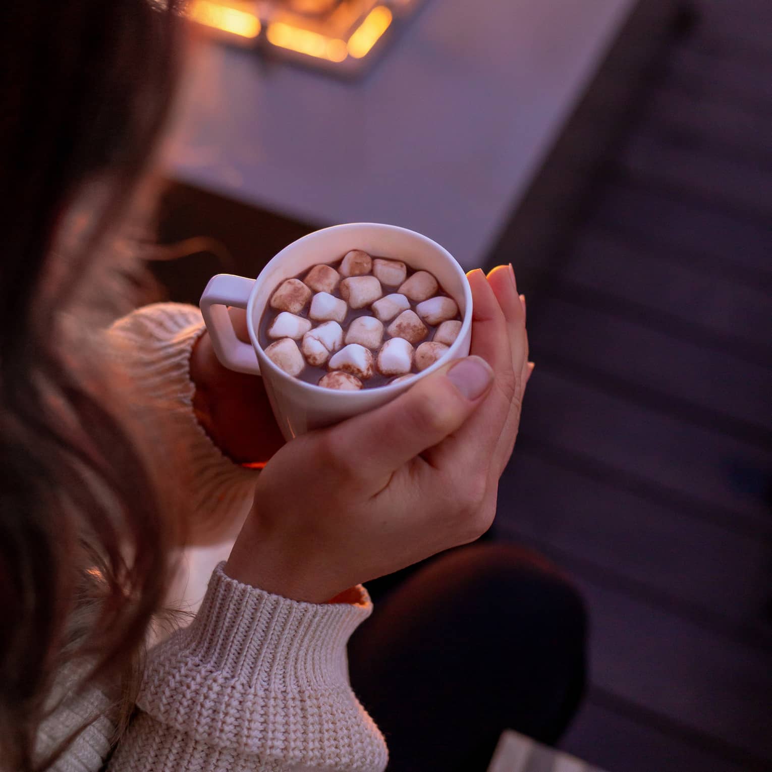 A woman holding hot chocolate by a fire.