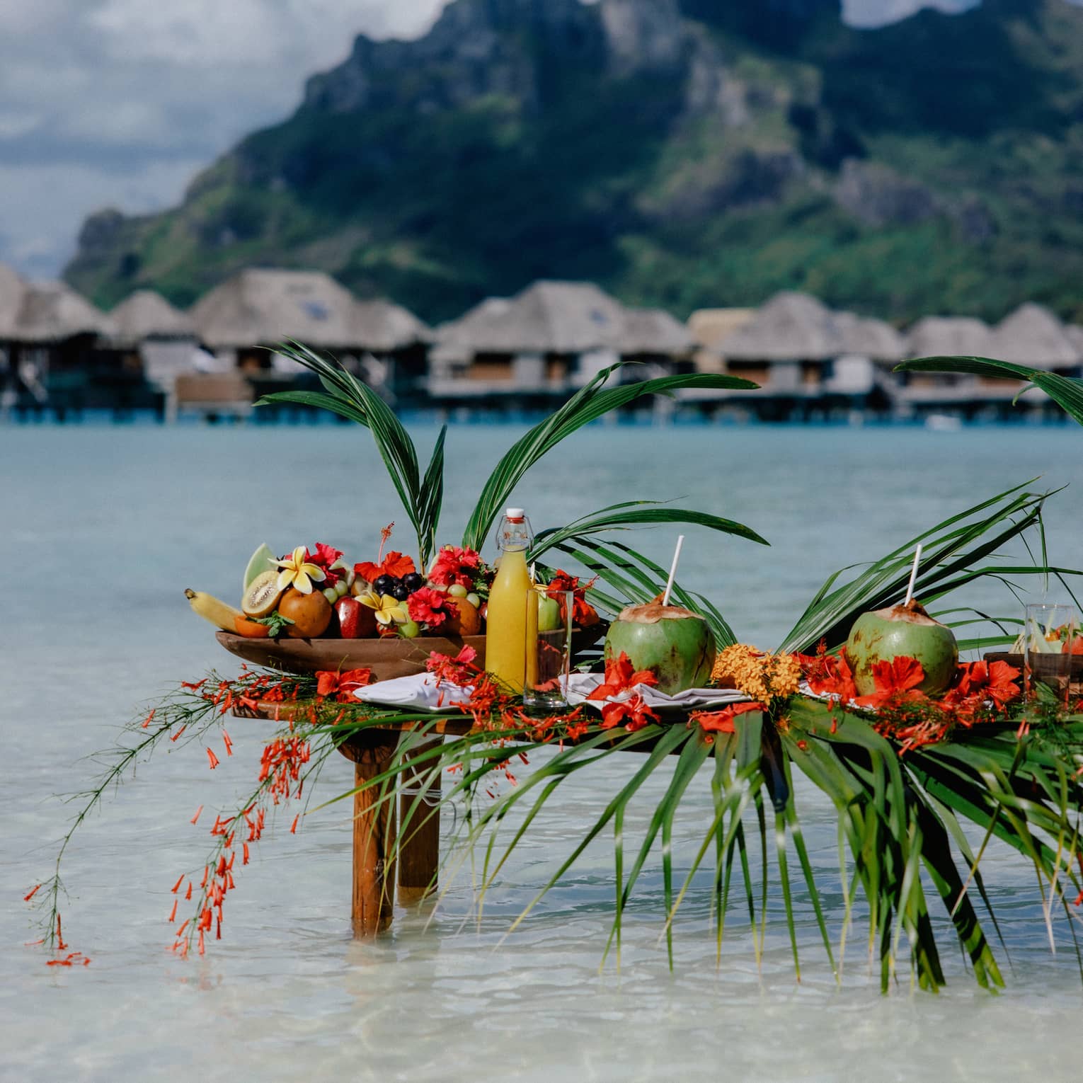 Small wooden table with legs submerged in clear water, topped with assorted fruit and coconuts with straws, mountain and overwater pavilions in background