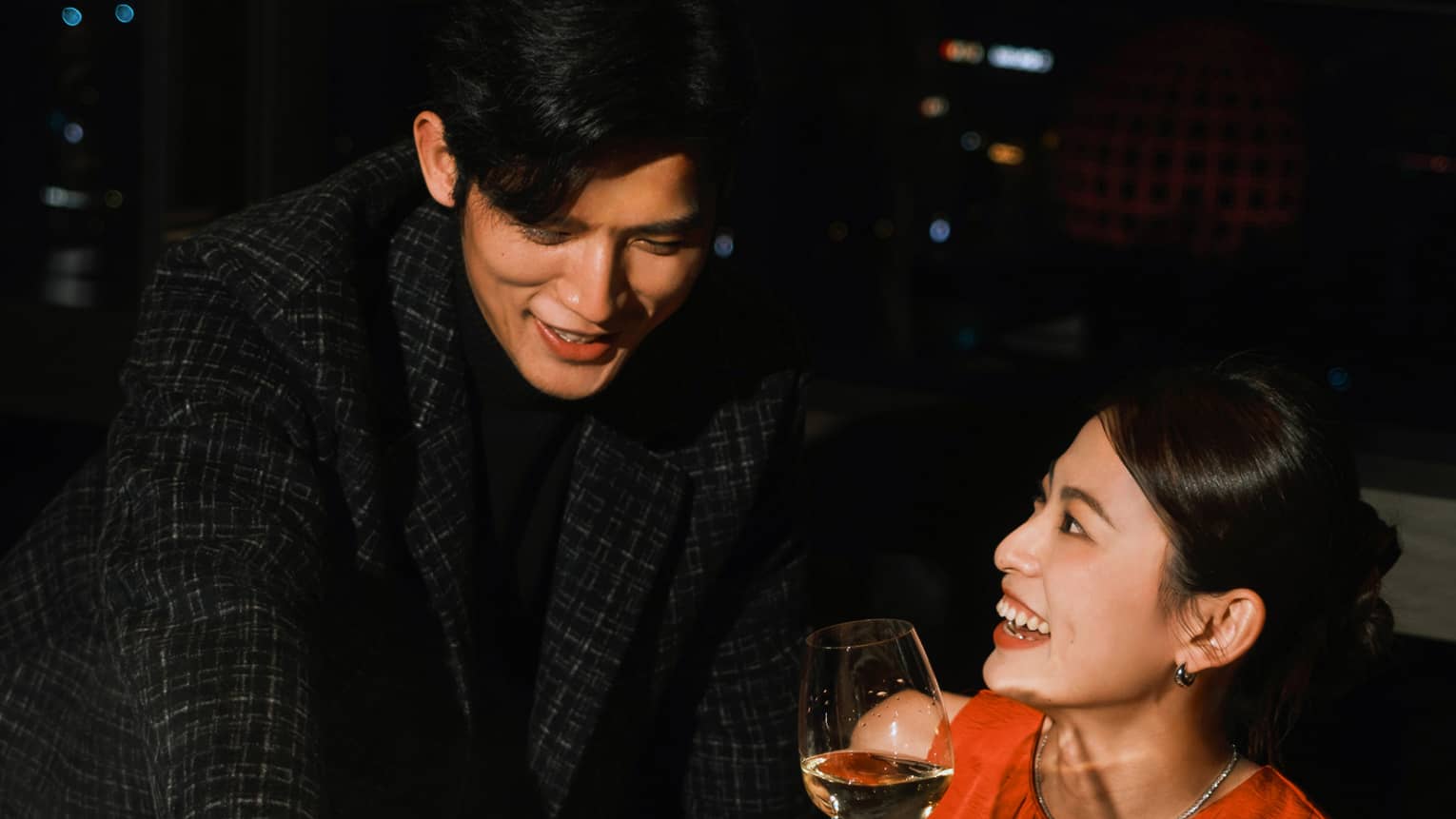 Guest reaches into a hot pot with chopsticks while another smiles holding a glass of white wine. Behind them is a night sky.