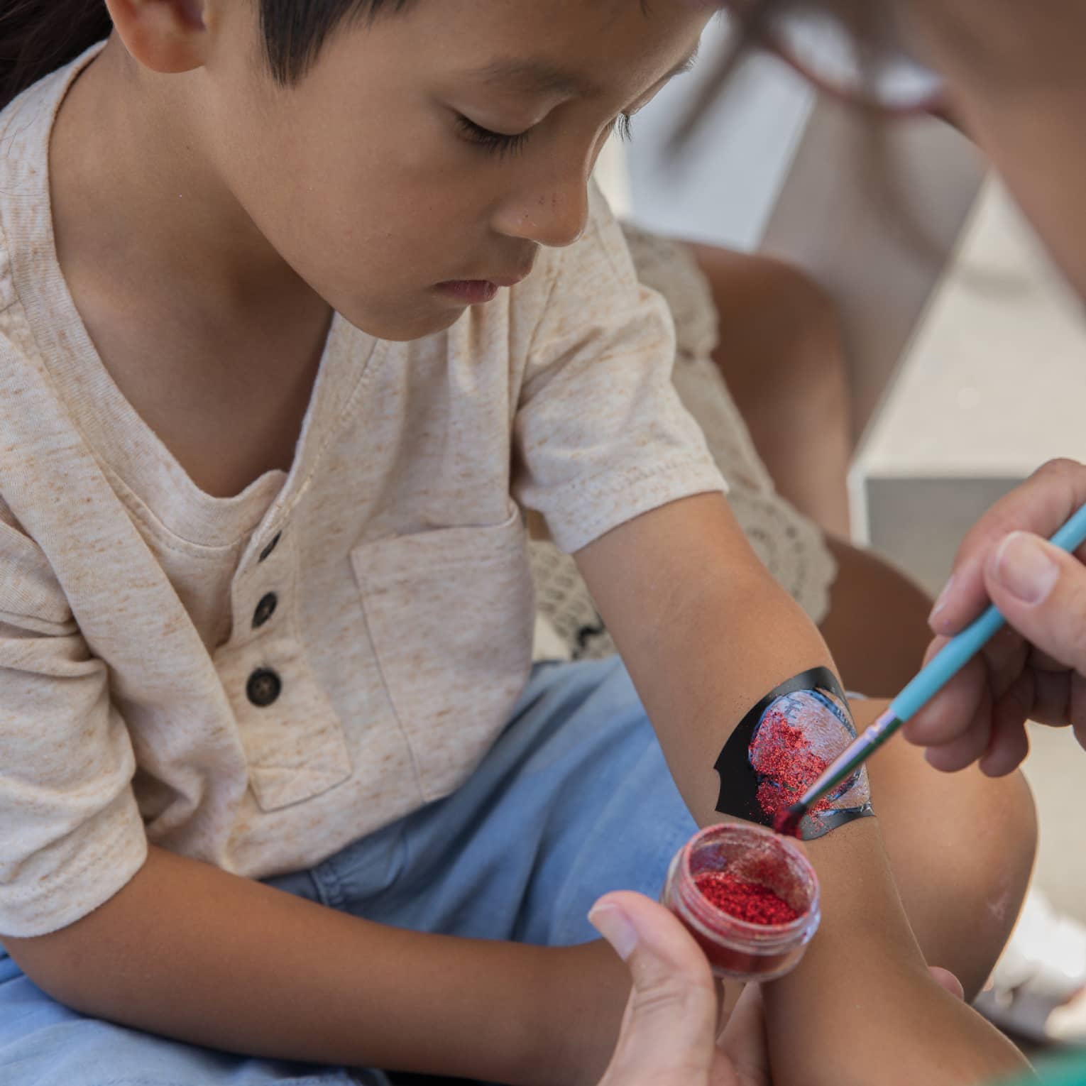 Child wearing beige shirt and light blue shorts sits getting their arm painted