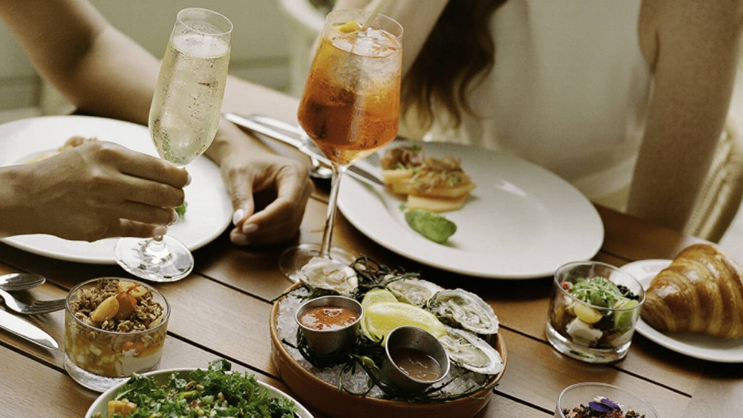Laughing guests enjoying cold beverages, raw oysters with sauces, smoked salmon, a kale salad, muesli and croissants.