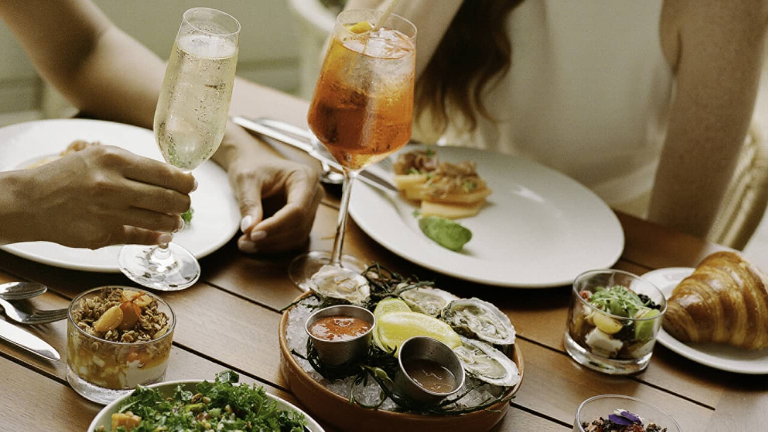 Laughing guests enjoying cold beverages, raw oysters with sauces, smoked salmon, a kale salad, muesli and croissants.
