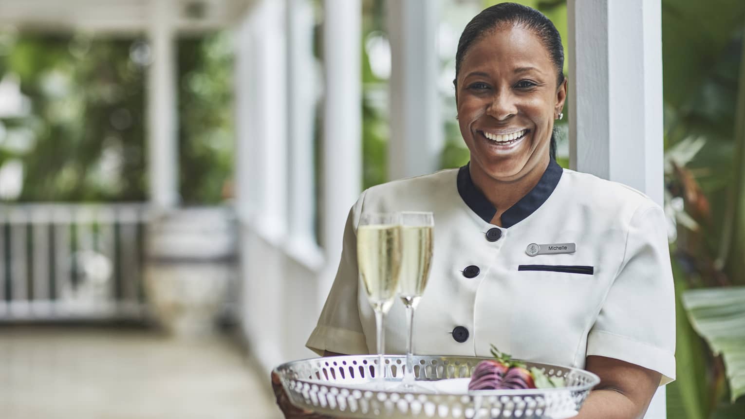 Smiling hotel staff holds tray with two glasses of Champagne on veranda