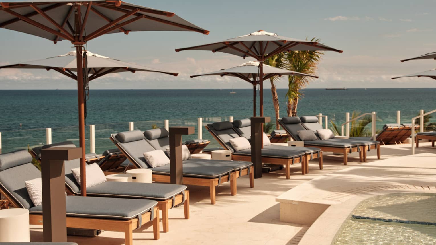 A curved pool deck with umbrellas and ocean view, featuring wooden lounge chairs with grey cushions