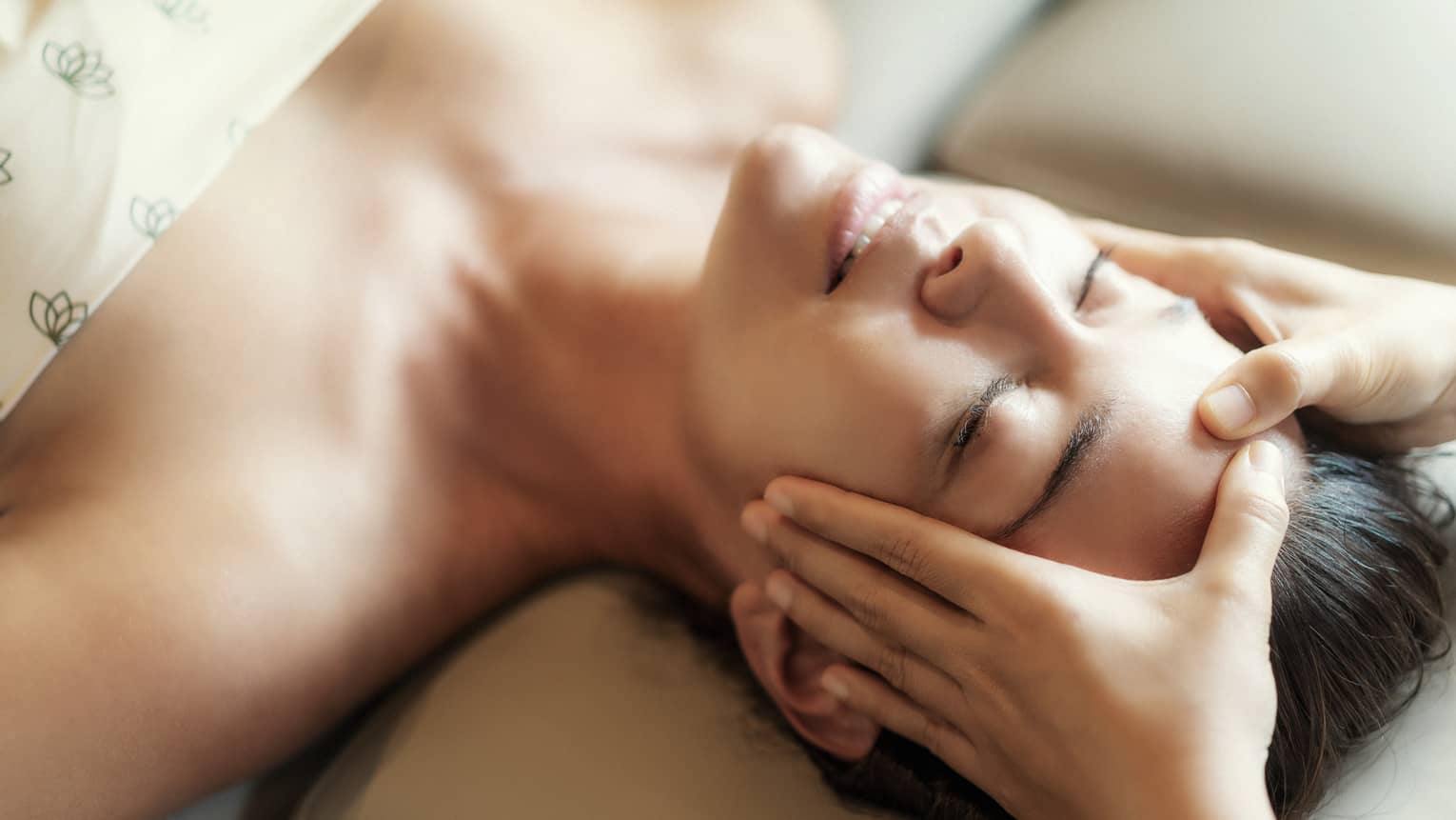 Spa facial, two hands rest on woman's forehead as she closes her eyes, lays under sheet on treatment table