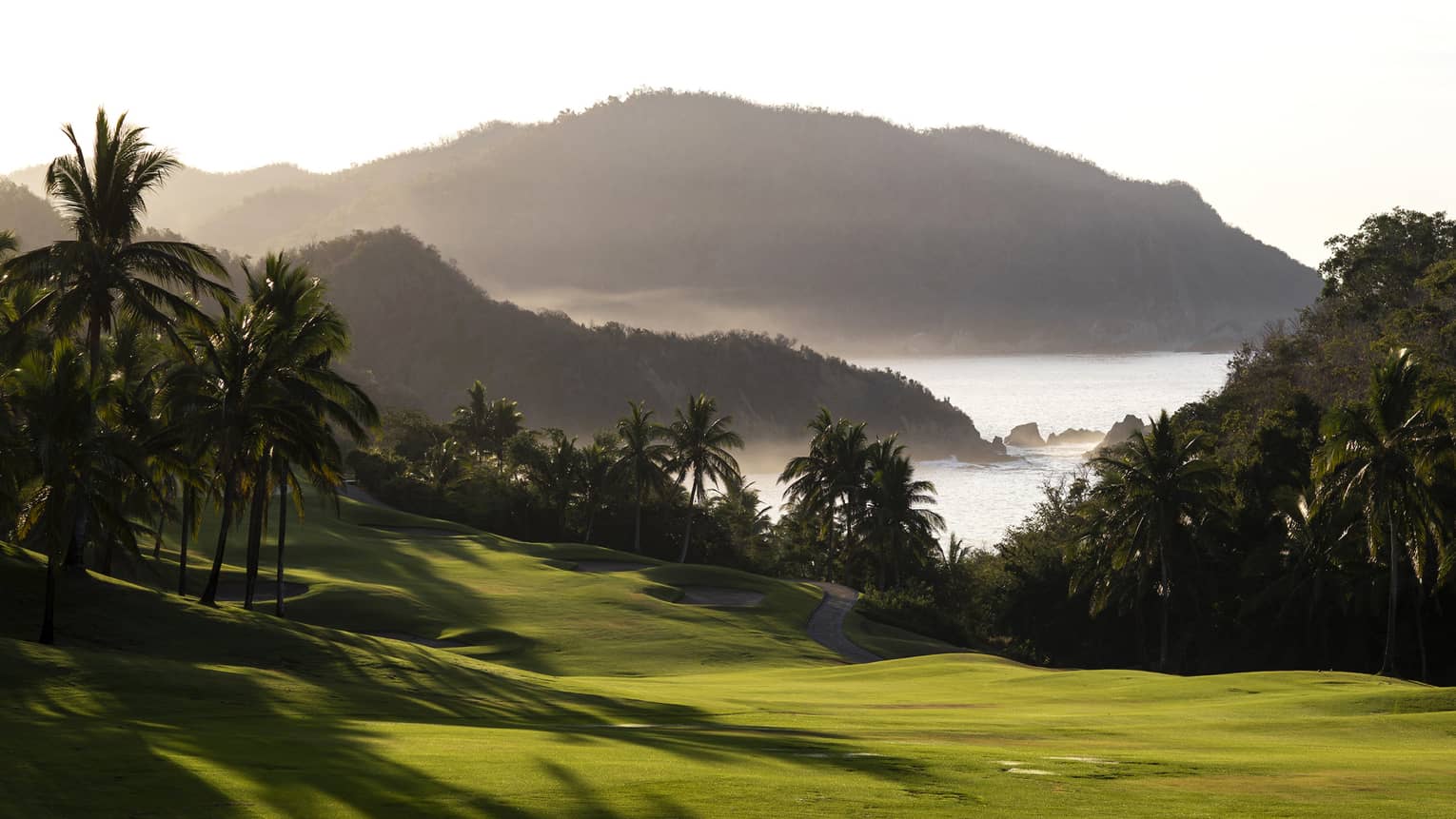 Palm trees cast long shadows on a majestic golf course against a backdrop of misty hills jutting out into the bay.
