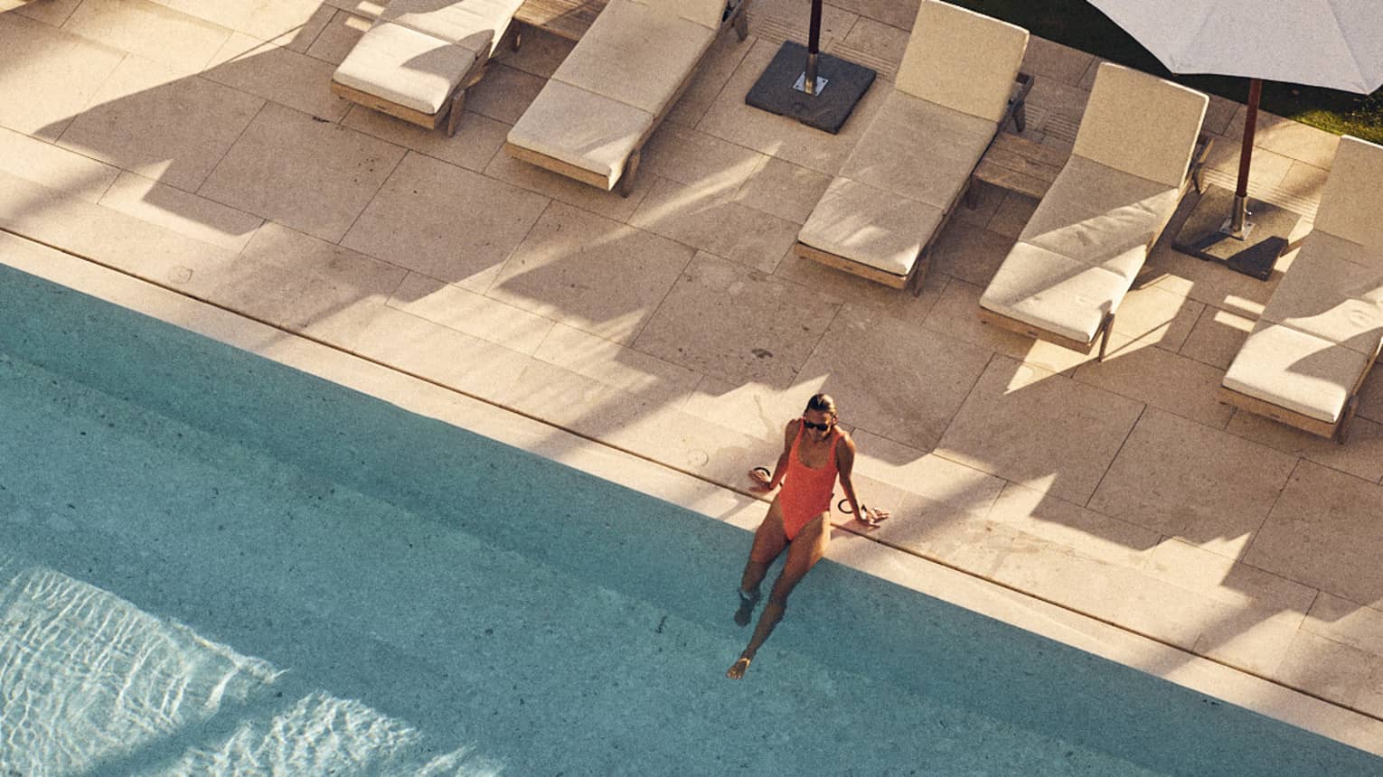 Aerial view of a woman in an orange swimsuit sitting on the edge of a large pool surrounded by lounge chairs and white umbrellas