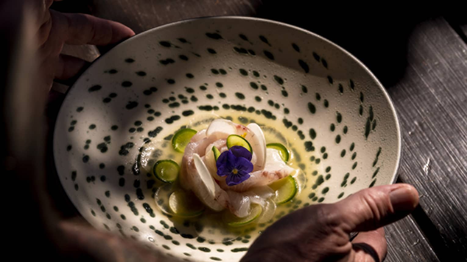 An artistically arranged seafood dish topped with a pansy flower is gently placed on a wooden table dappled with light.