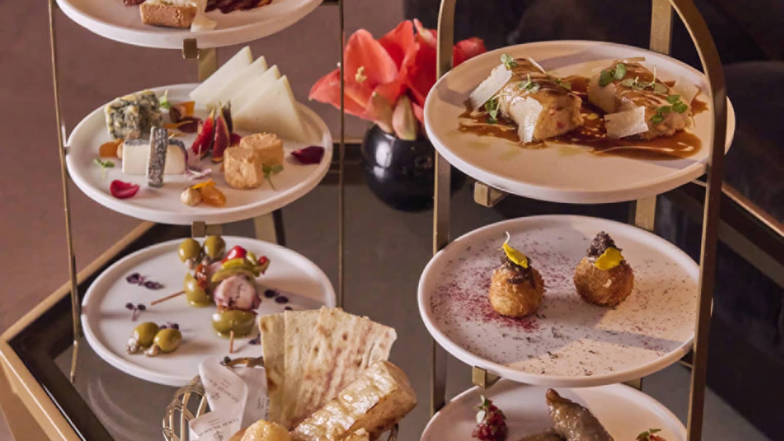 Tiered white and gold trays with a variety of savoury food of meats, cheese, olives, bread and pastries laid out on a table.