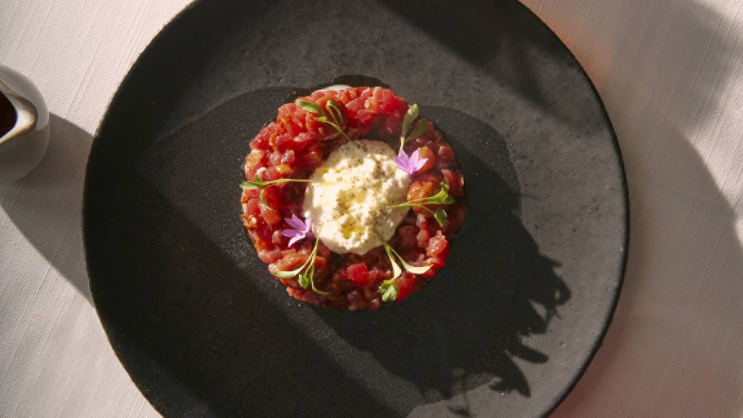 Round Beef Tartare in centre of black plate with two cocktails beside it