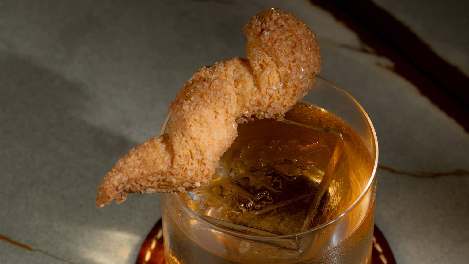 An amber-hued drink in a clear tumbler with a tan garnish resting on the rim
