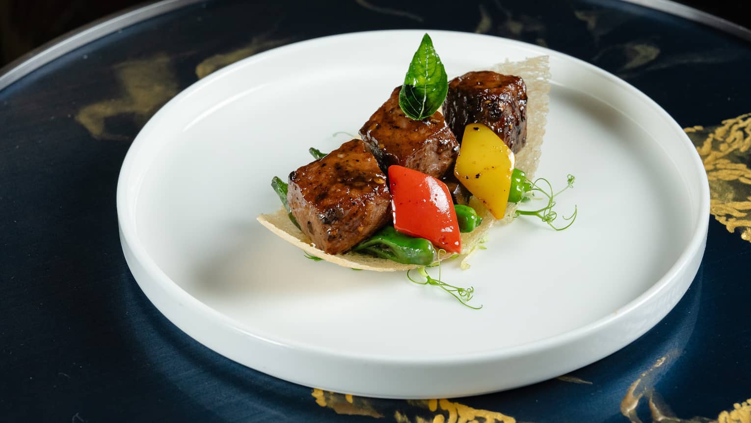 Three cubes of grilled meat with diamond-shaped green and red pepper slices and microgreens, cradled by a curved rice wafer.