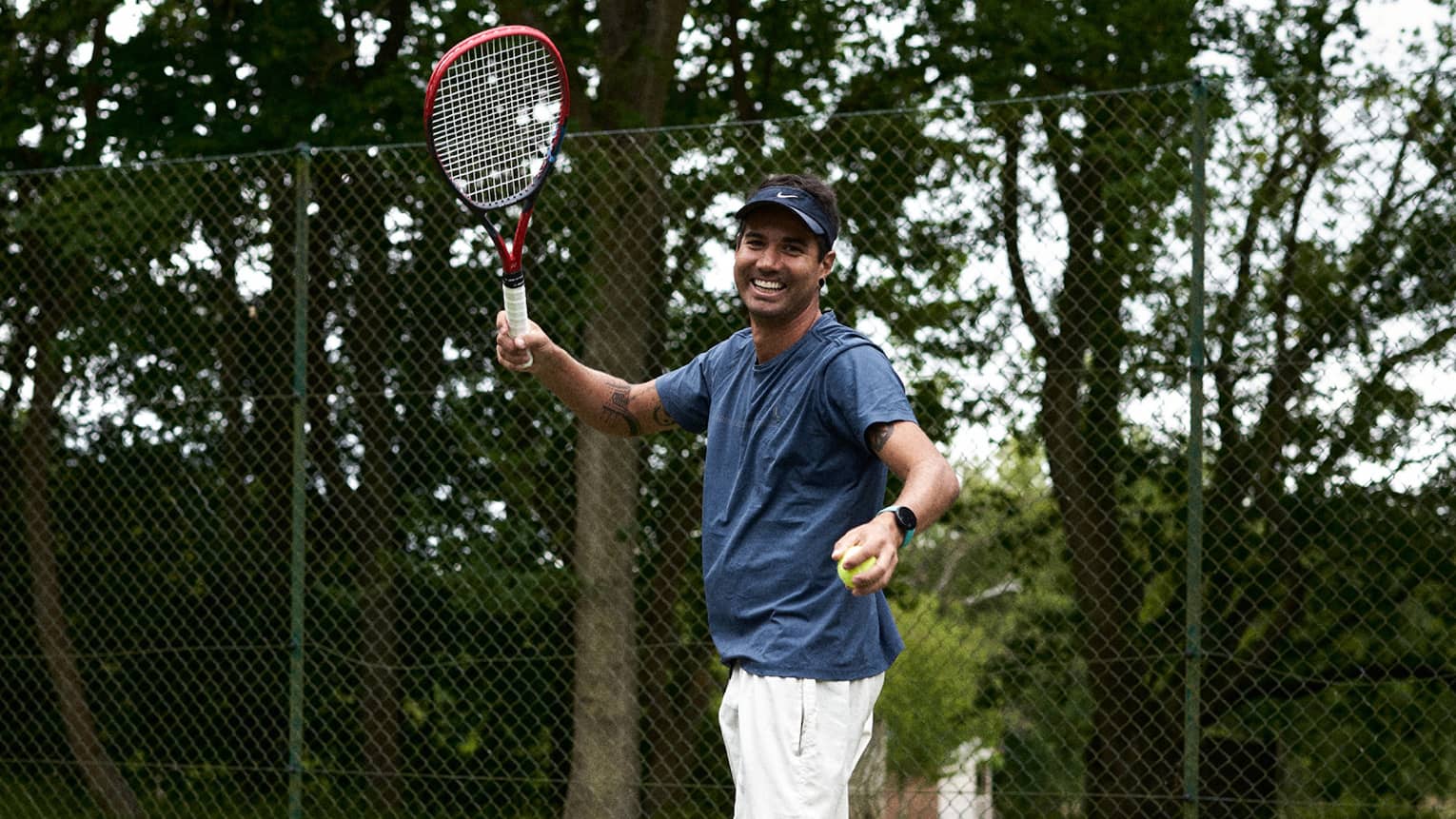Person standing and smiling while readying to serve on green tennis court with fence and trees in backdrop