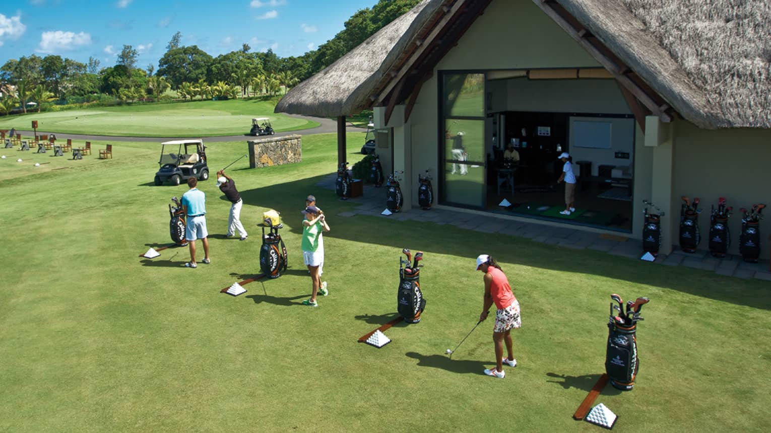 Row of women and men swinging golf clubs on driving range in front of clubhouse 