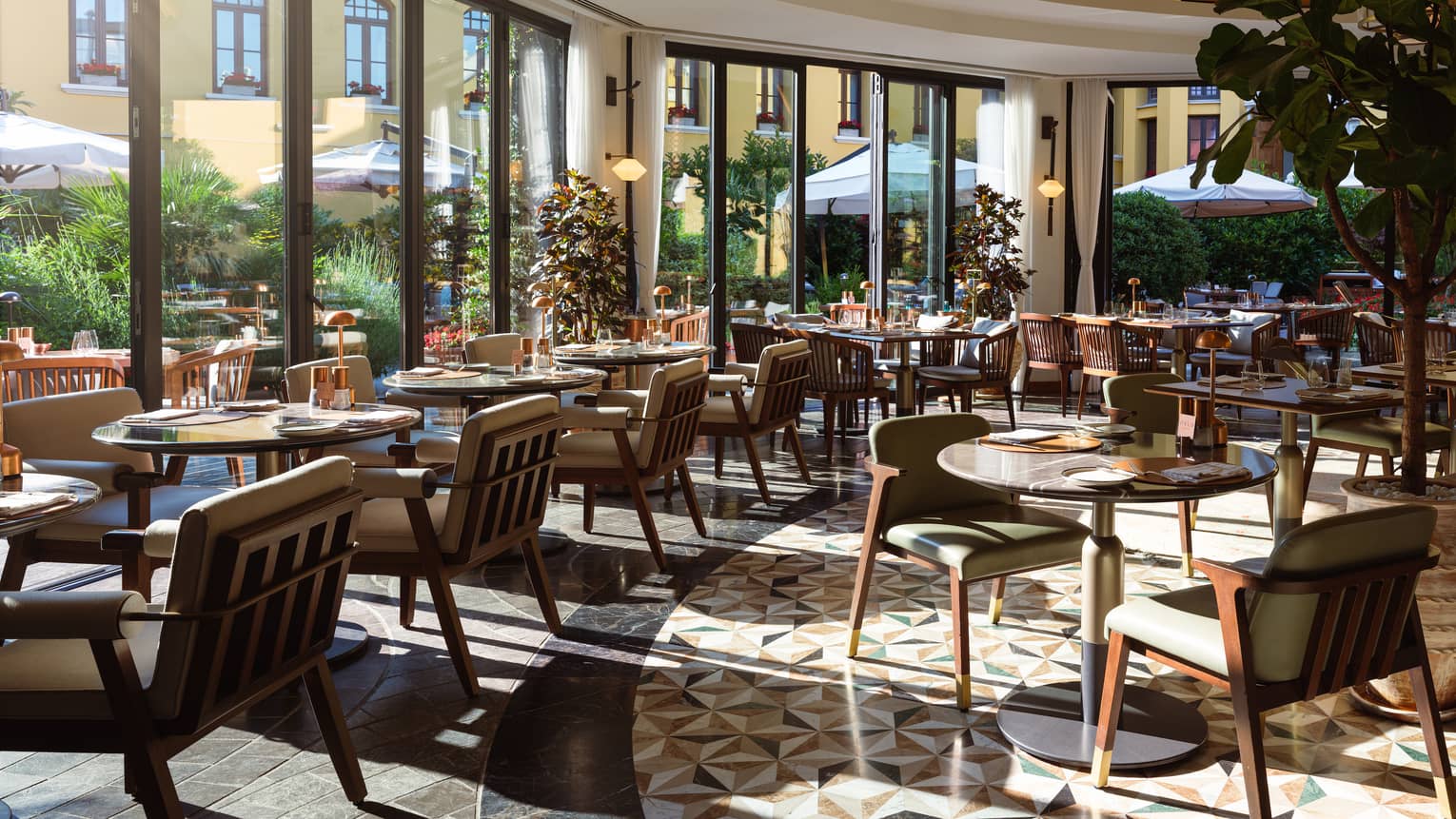 A light-filled indoor dining space with floor-to-ceiling windows along a curved wall, letting in sunny views of the outdoor dining area. Small tables with upholstered seating fill the space.