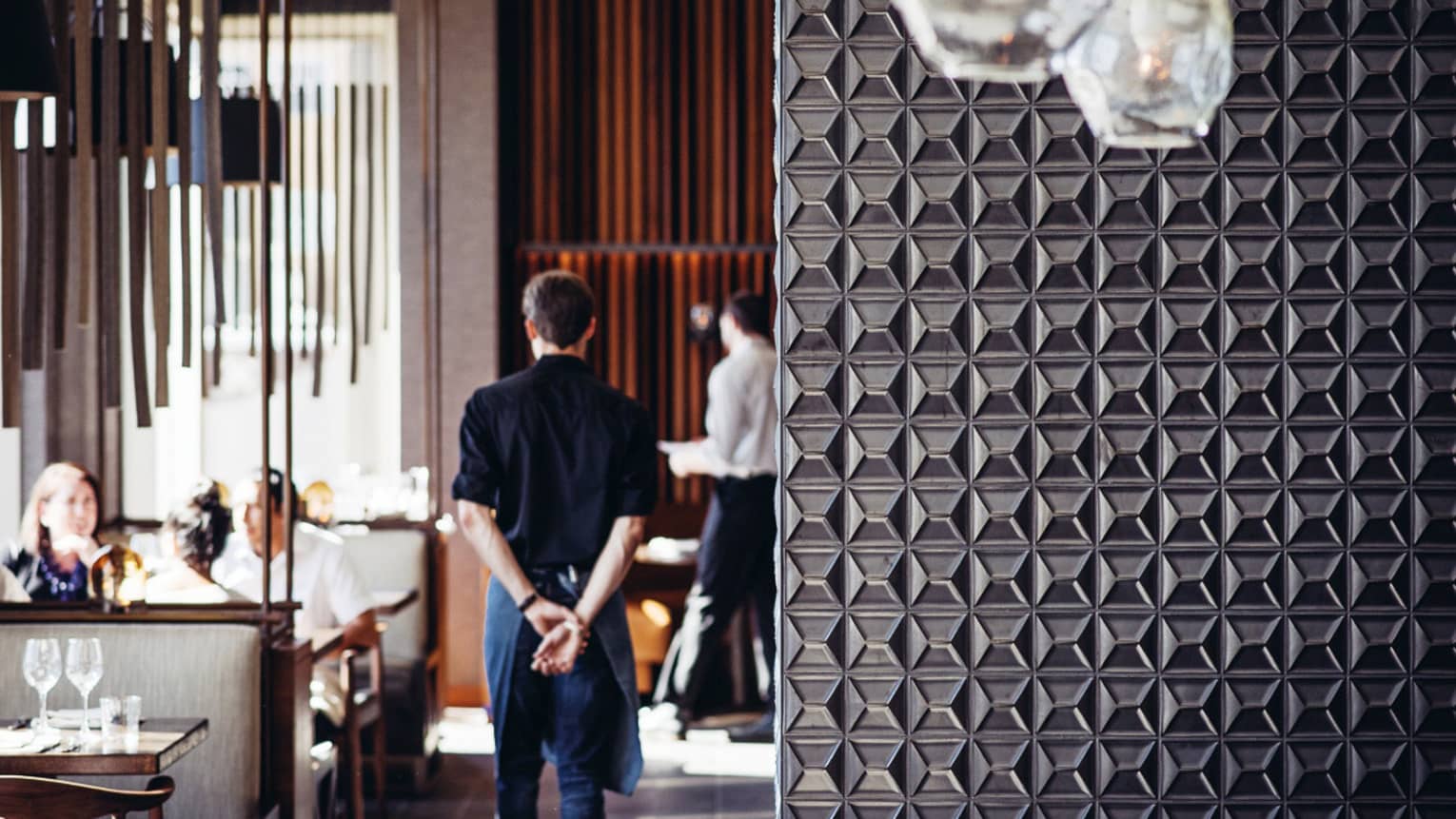 Server walks with hands behind back past dining tables, beyond decorative metal wall