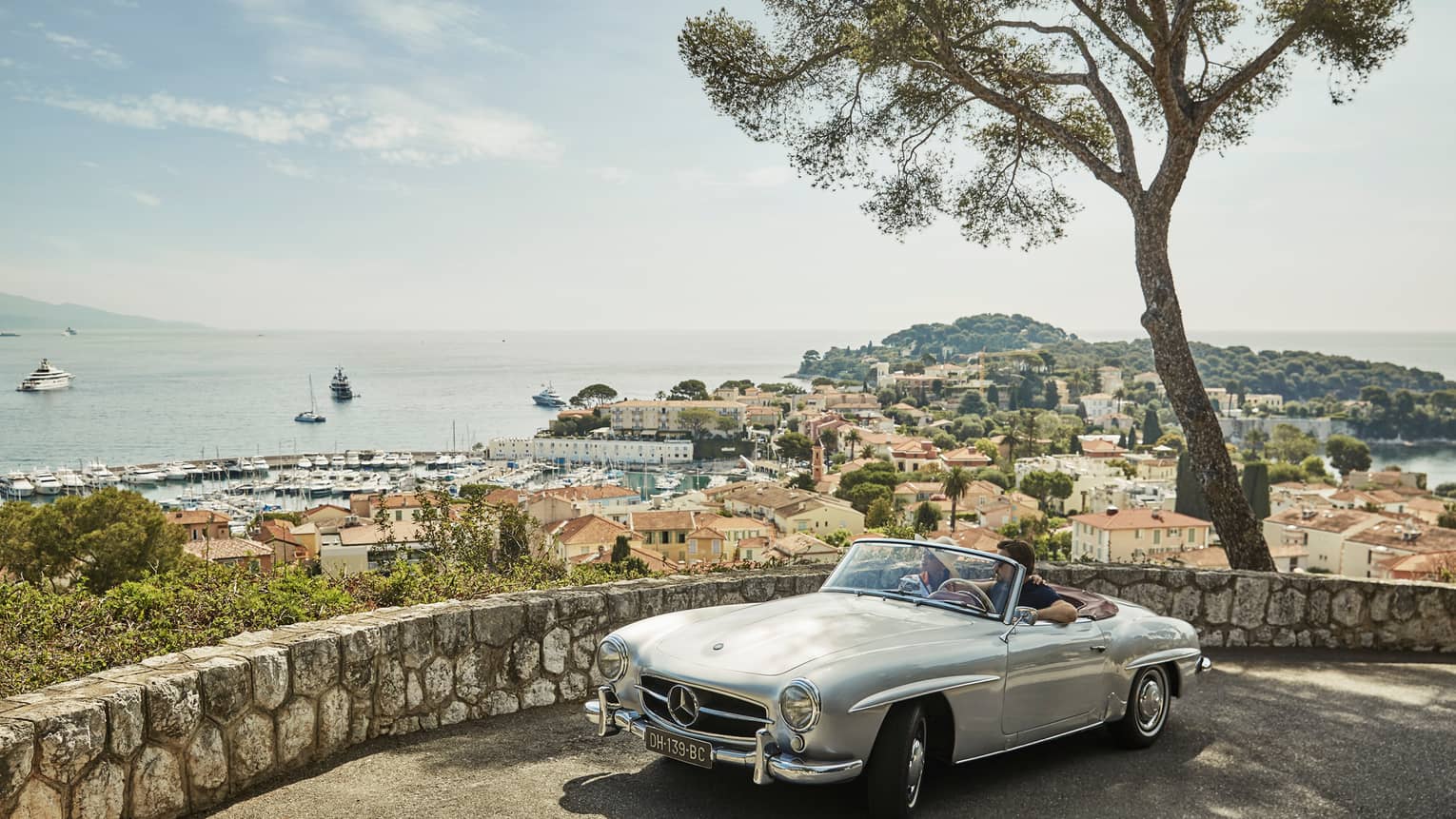 Couple in luxury vintage white car parked under tree by stone wall overlooking red roofs, water