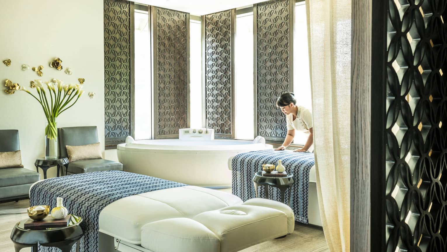 Large white spa table in front of white spa tub, silver wall panels, woman places blanket on table