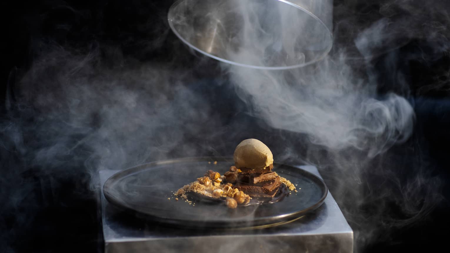 Food on a black tray with glass cover being lifted off of it, with wisps of smoke moving around the food.