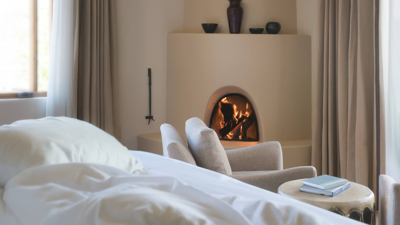 Hotel bed pillows, white armchair in front of curved clay fireplace, vases on mantle