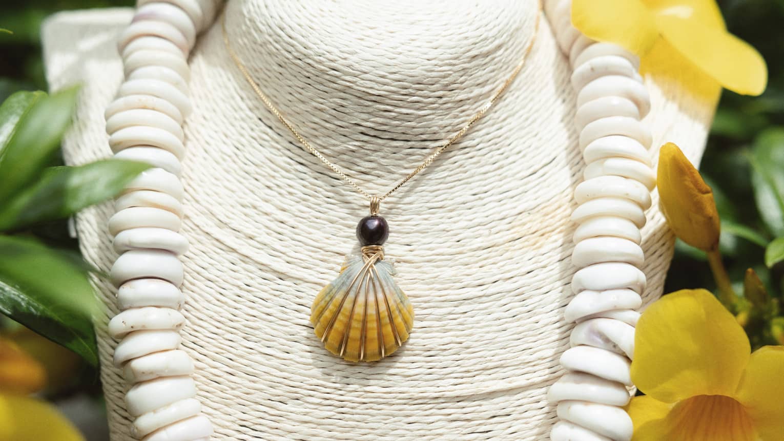 A jewelry display of two shell necklaces, with greenery and yellow flowers surrounding it and in the background