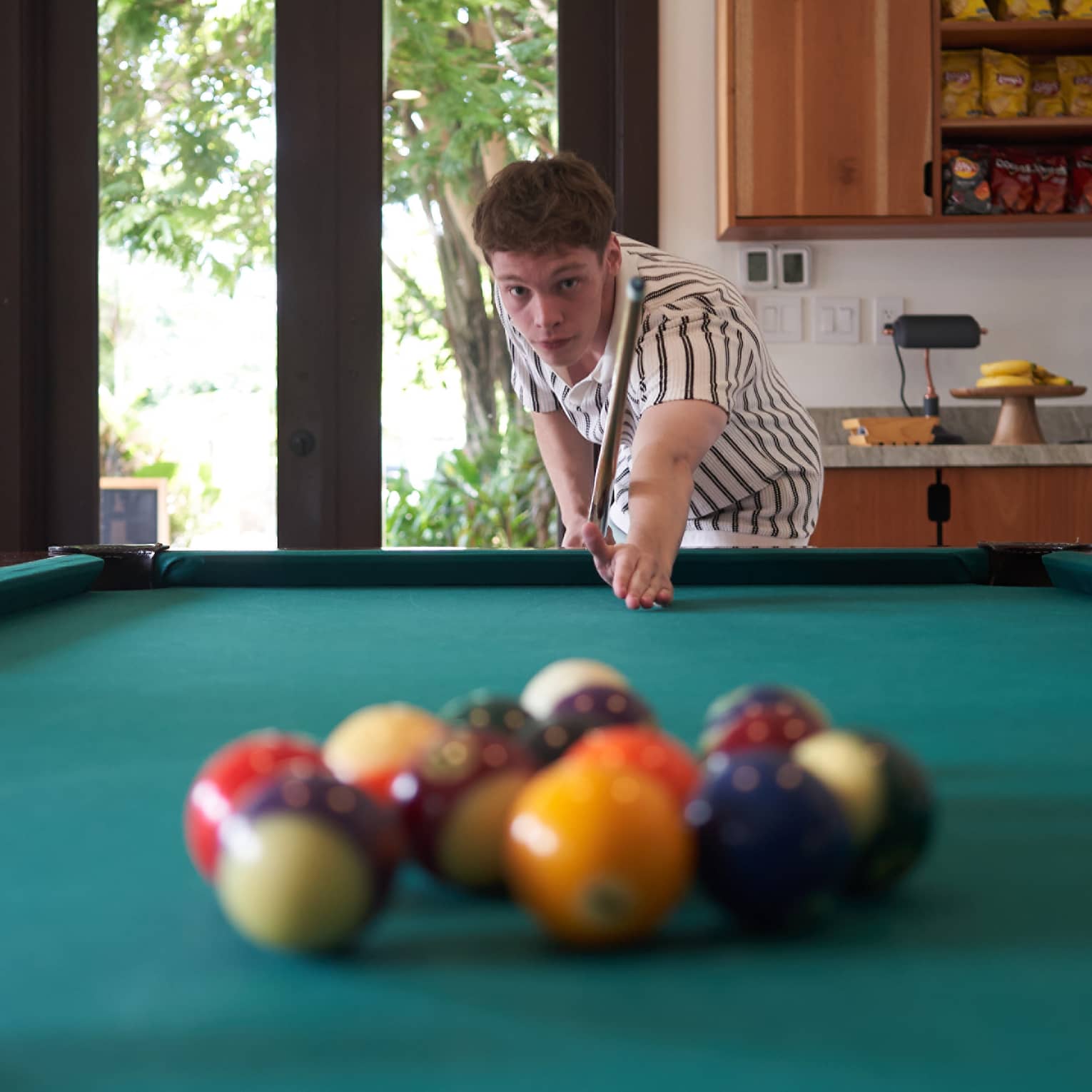 Teen wearing green-and-white button down short-sleeve shirt aims a cue at a collection of billiard balls on a green pool table