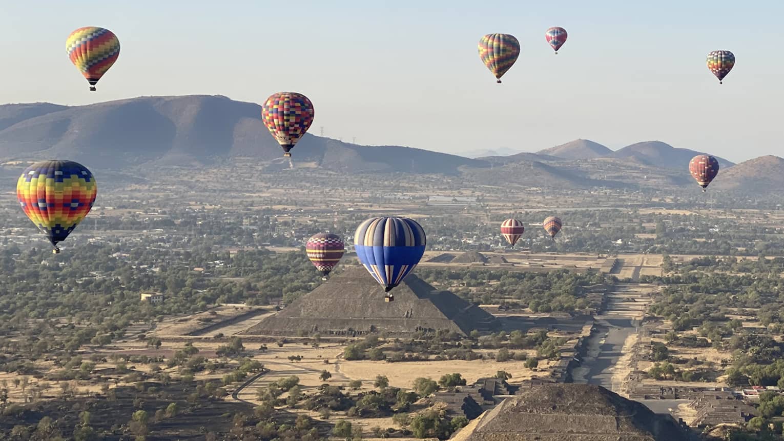 Hot-air balloons floating over old temples in a dessert covered in shrubs.