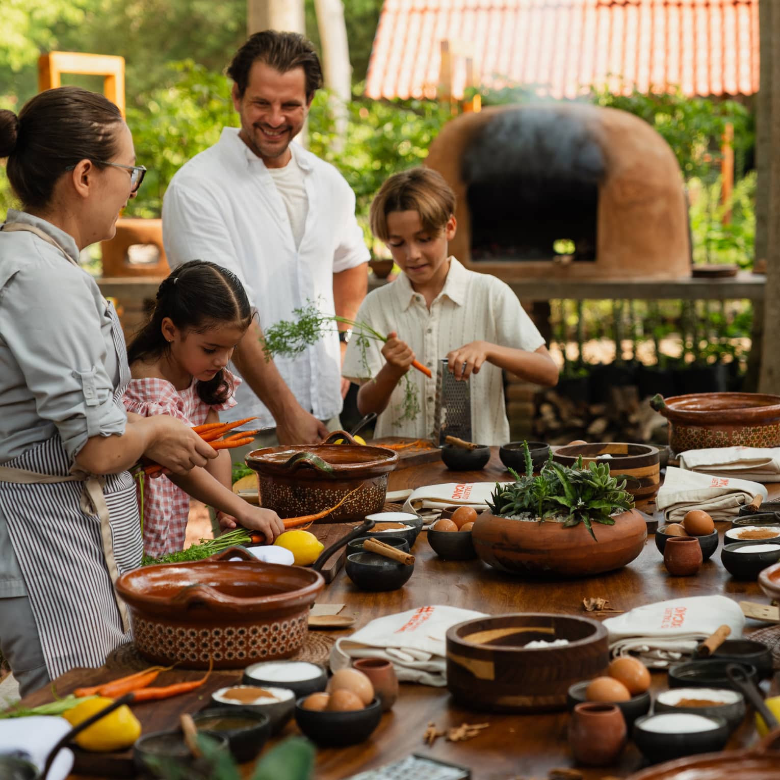 Adults and children standing around an outdoor dining table with various dishes and utensils on it, while an outdoor oven runs in the background.